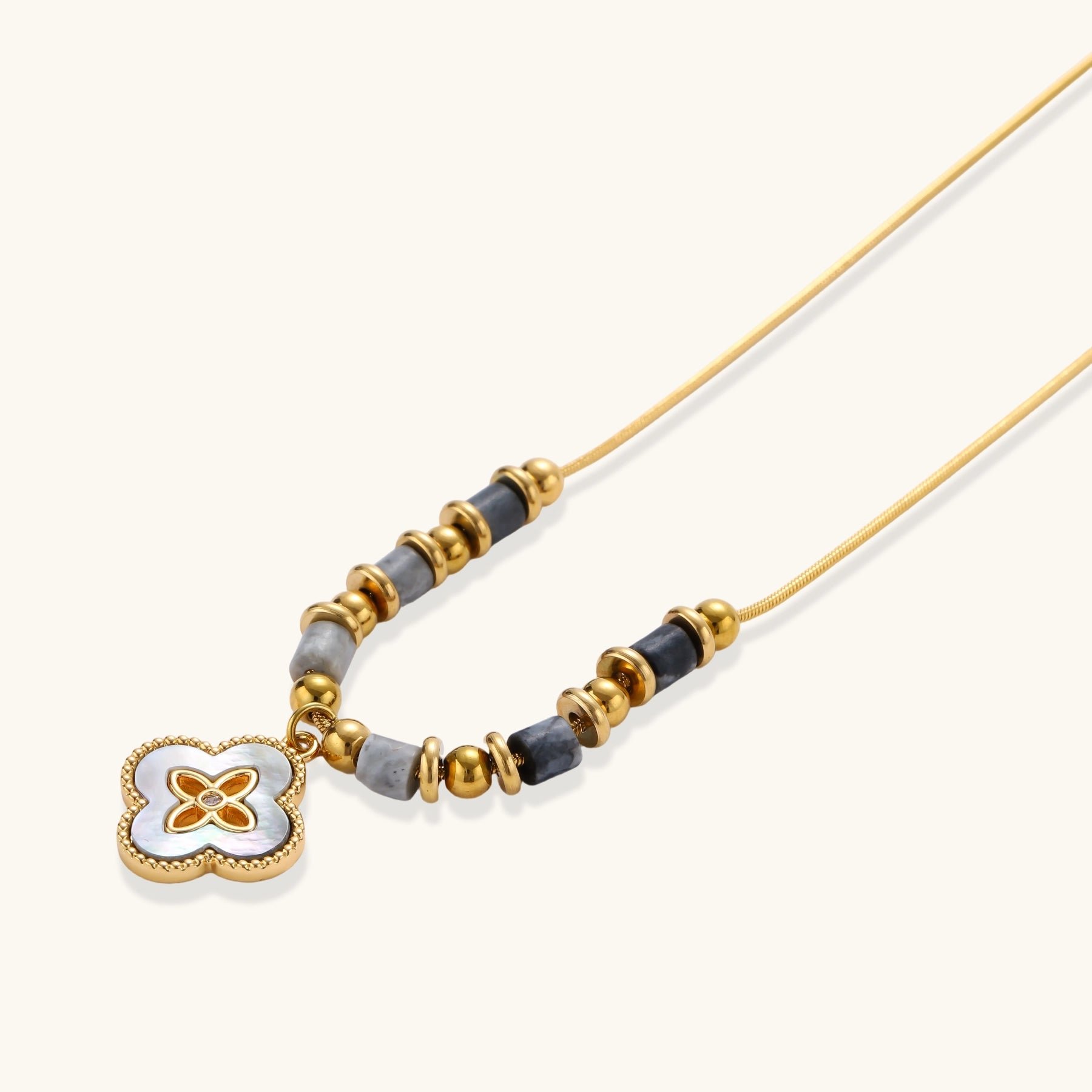 Gold-toned beaded necklace with grey stones and quatrefoil abalone shell pendant on cream background.