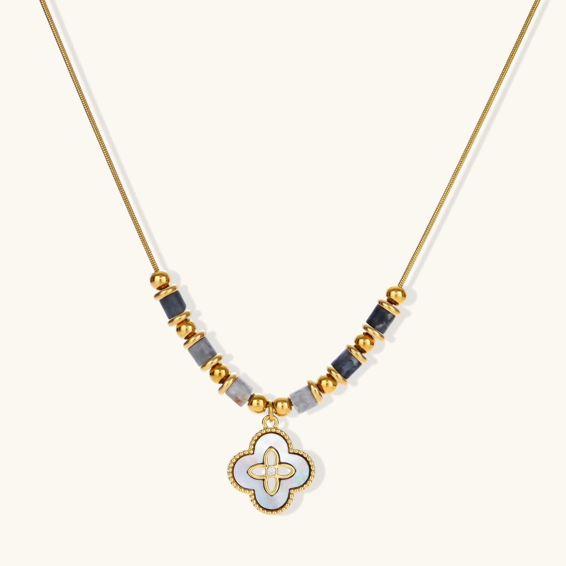 Gold-toned beaded necklace with grey stones and quatrefoil abalone shell pendant on cream background.