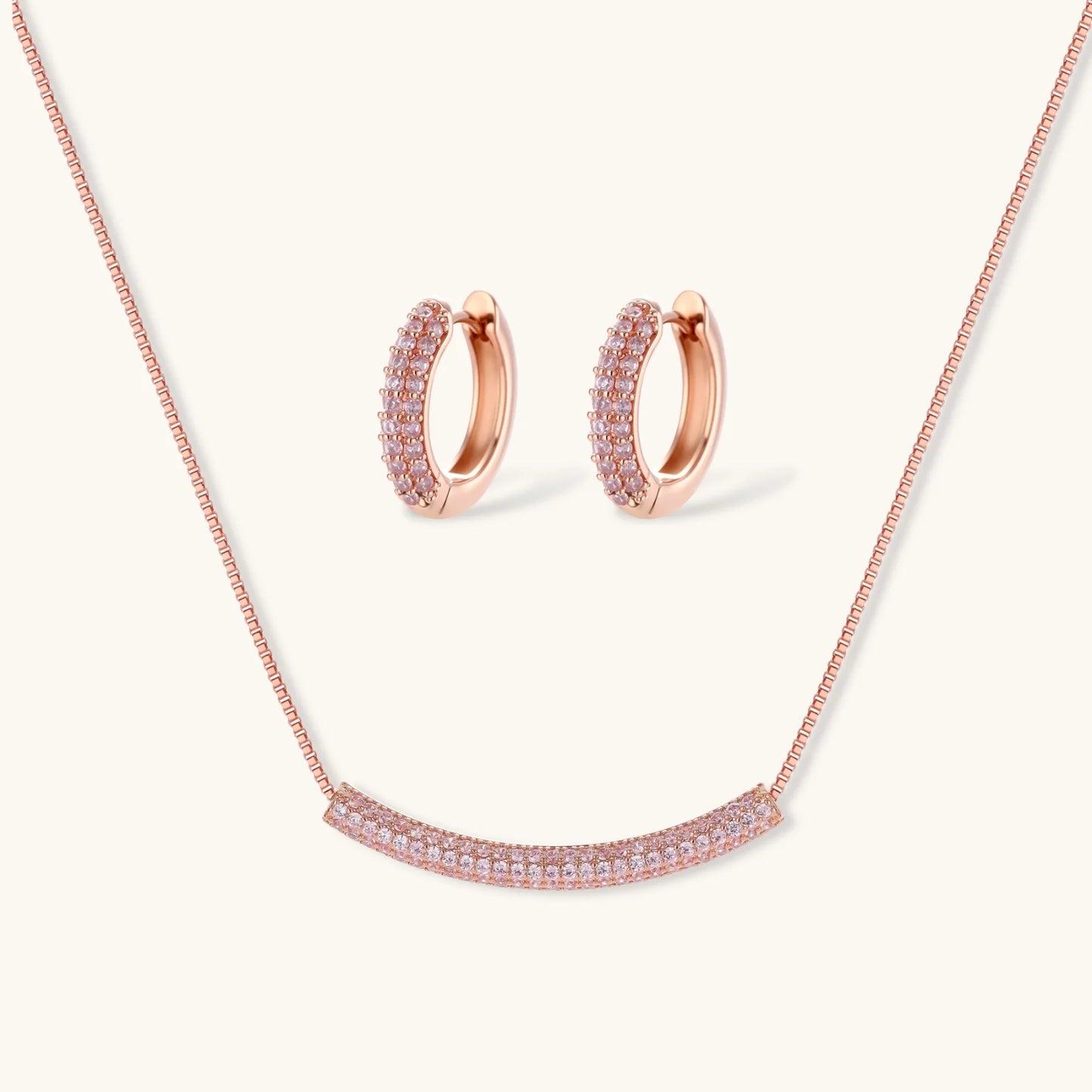 Rose gold jewelry set with pavé cubic zirconia stones including curved necklace, hoop earrings, and adjustable ring on cream background.